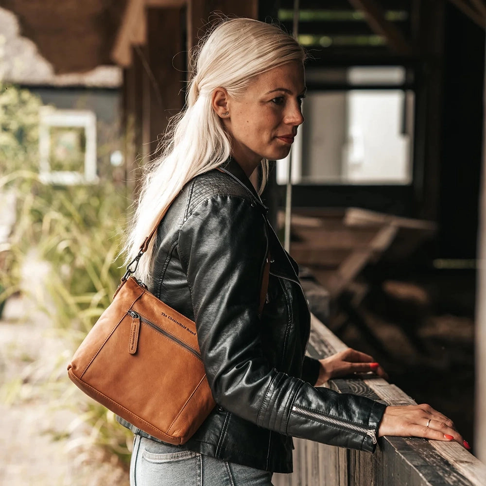 blond kvinna i svart skinnjacka lutar sig  mot staket med cognacsfärgad läderväska över axeln av modellen Faro från chesterfield brand